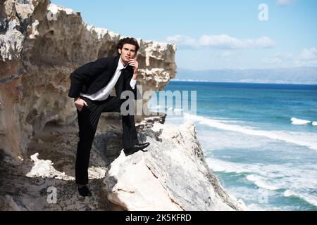 Was für eine schöne Aussicht ... ein hübscher Geschäftsmann, der für die Kamera auf einem Felsen mit Blick auf den Ozean posiert. Stockfoto