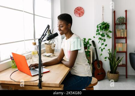 Lächelnde junge Podcasterin, die Radio-Podcast aus dem modernen Heimstudio aufnimmt Stockfoto