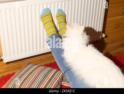 Frau und ihre weiße Katze sitzen in der Nähe der Zentralheizungsbatterie. Sie wärmt seine Füße in gestreiften Socken auf der Batterie. Sanktionen, Russland. Konzept Wintersaison Stockfoto