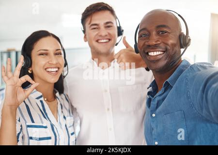 Frau, Männer und Selfie von Callcenter-Agenten mit Headset, Daumen hoch und ok. Teamarbeit, Unterstützung und Hilfe, freundliches, begeistertes Telemarketing- oder crm-Team Stockfoto
