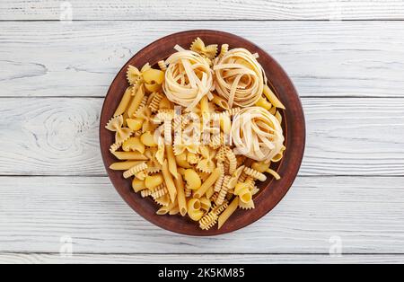 Verschiedene Arten und Formen italienischer Pasta in Keramikplatte auf weißem Holzhintergrund Stockfoto