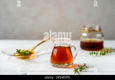 Thymianhonig in kleinem Krug und Glas, kleiner Thymianzweig auf weißem Holztisch mit grauem Hintergrund Stockfoto