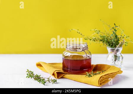 Thymianhonig im kleinen Glas, frischer Thymian, gelbe Serviette auf weißem Tisch mit gelbem Hintergrund Stockfoto