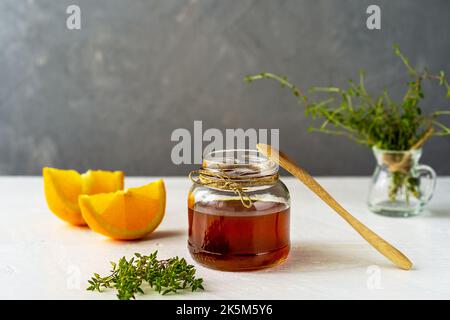 Thymianhonig im kleinen Glas, Teelöffel aus Holz, kleiner Thymianzweig, orangefarbene Scheiben auf weißem Tisch mit grauem Hintergrund Stockfoto