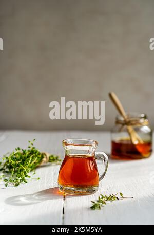 Thymianhonig in kleinem Krug und Glas, kleiner Thymianzweig auf weißem Holztisch mit grauem Hintergrund Stockfoto