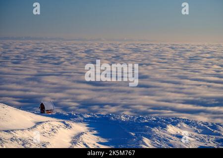 Eine Luftaufnahme aus weißen Wolken über Bergen und ein kleines Haus auf einem verschneiten Hang im Kopaonik-Berg, Serbien Stockfoto