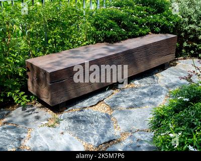 Leerer Sitz aus einem großen Holzbalken in rechteckiger Form, der auf dem Steinboden im grünen Garten mit Baumschatten an sonnigen Tagen aufgestellt wurde. Stockfoto