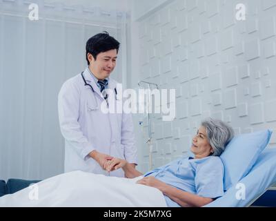 Der freundliche asiatische männliche Arzt in weißem Anzug besucht, spricht und unterstützt, indem er die Hand eines glücklichen älteren Patienten in hellblauem Kleid in der Hand hält Stockfoto