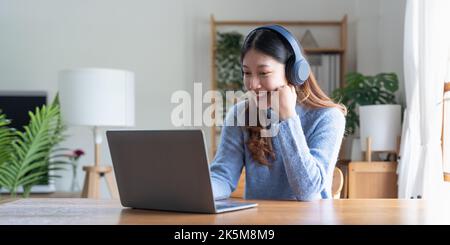 Junge asiatische Frau spricht bei einem Videoanruf am Tisch mit Kopfhörern. Online-Fernarbeit oder Lernkonzept Stockfoto