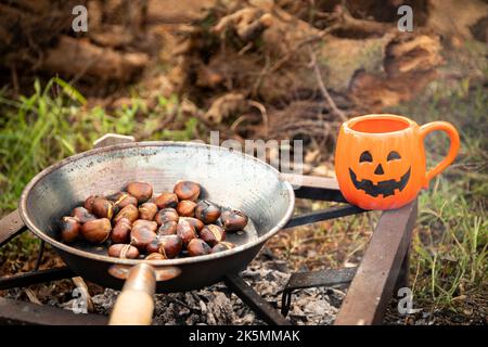 Rösten von Kastanien in einer Pfanne auf heißen Kohlen im Freien und Kürbis-halloween-Becher. Lagerfeuer Kochen im Herbst, halloween Camping Inspiration. Stockfoto