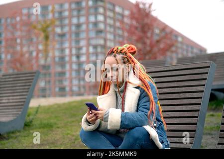 Junge Frau mit Dreadlocks in warmer Kleidung, die im Stadtpark auf einer hölzernen Sonnenliege auf dem Mobiltelefon surft. Sorglose Hündin mit farbenfroher Frisur usi Stockfoto