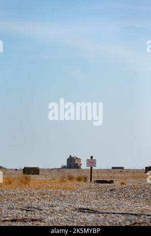 Flache Schindellandschaft mit Warnschild und Gebäuden in der Ferne, Orford Ness, Suffolk, England Stockfoto