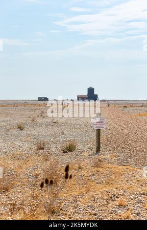 Flache Schindellandschaft mit Warnschild und Gebäuden in der Ferne, Orford Ness, Suffolk, England Stockfoto