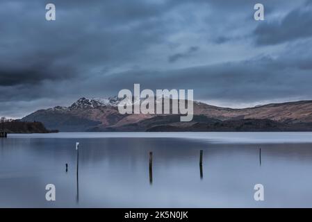 Atemberaubendes Landschaftsbild von Loch Lomond und schneebedeckter Bergkette in der Ferne vom kleinen Dorf Luss aus gesehen Stockfoto