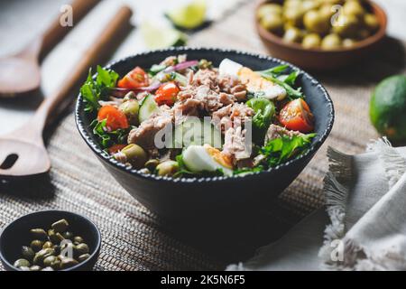 Thunfischkonserven-Salat mit frischem Gemüse, Kapern und Oliven in einer schwarzen Schüssel. Gesundes Mittag- oder Abendessen. Stockfoto