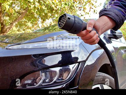 Hand hält ein Elektroauto-Ladekabel mit schwarzem Elektroauto hinter sich Stockfoto