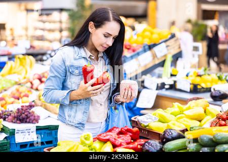 Frau, die Gemüse kauft, hält auf dem Bauernmarkt rote Paprika und ihren Geldbeutel in den Händen. Stockfoto