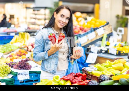 Eine schöne lächelnde Frau wählt zwei rote Paprika aus einer großen Auswahl an frischem Obst und Gemüse auf dem Markt aus. Stockfoto