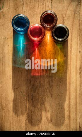 Vier Glasgefäße auf einem alten Holztisch, die Wasser mit Farbe enthalten und das Licht reflektiert die Farbe auf dem Holz Stockfoto