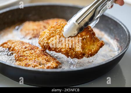 Detail einer Zange, die gebratenes Schnitzel aus einer brutzelnden Pfanne nimmt. Stockfoto