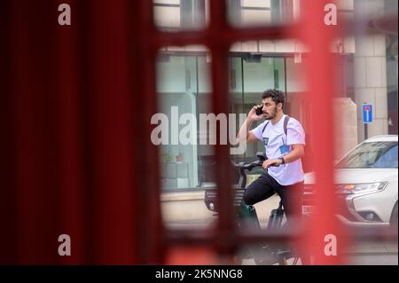 LONDON - 21. Mai 2022: Mann nutzt Mobiltelefon, während er ein Elektrofahrrad ausleih. Über die herkömmliche Telefonbox gesehen Stockfoto