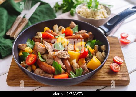 Gebratene Würstchen, große Stücke Schweinefleisch, mit gelben Paprika, Kirschtomatenscheiben, Petersilienblättern und Couscous. Ein buntes Gericht in einer Pfanne. Stockfoto