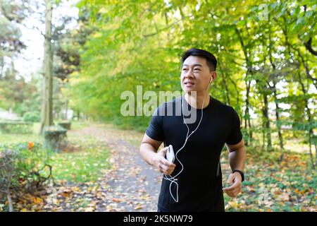 Mann beim Joggen am Morgen im Herbstpark, asiatischer Sportler in Sportkleidung mit Kopfhörer und Telefon im Wald hört Musik beim Joggen und Spazierengehen aktiven Lebensstil Stockfoto