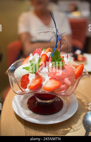 Köstliche Tasse mit Eis, Erdbeeren und Schlagsahne. Junge unkenntliche Frau im Hintergrund Stockfoto