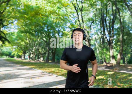 Mann beim Joggen am Morgen im Herbstpark, asiatischer Sportler in Sportkleidung mit Kopfhörer und Telefon im Wald hört Musik beim Joggen und Spazierengehen aktiven Lebensstil Stockfoto
