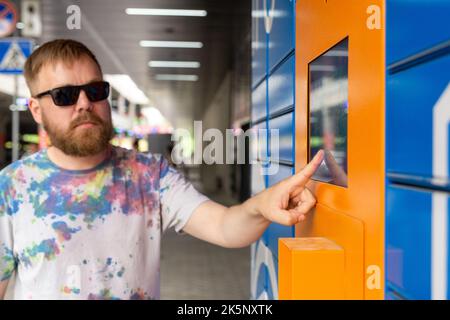 Der Mann gibt den Paketcode auf dem Bildschirm ein und holt das Paket vom automatischen Postautomaten ab. Mann hält Smartphone über automatische Self-Service-Mail-Terminal cl Stockfoto