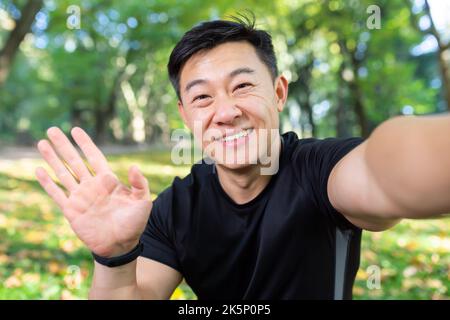 Asiatischer Sportler Läufer im Herbstpark, der die Smartphone-Kamera anschaut und lächelt, mit Freunden im Videoanruf spricht, Mann in Sportkleidung für Fitness im Herbstwald. Stockfoto
