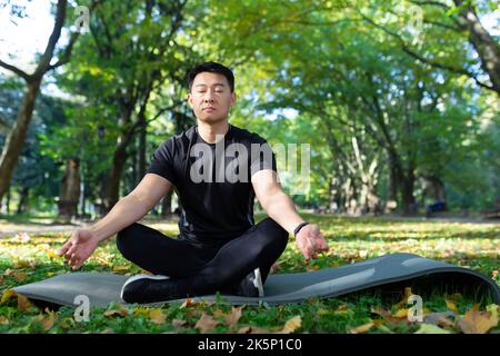 Porträt eines asiatischen Mannes im Herbstpark, der in Lotusposition auf einer Fitnessmatte sitzt, einen Sportler, der sich nach dem Training mit Atemübungen ausruhte, Stockfoto