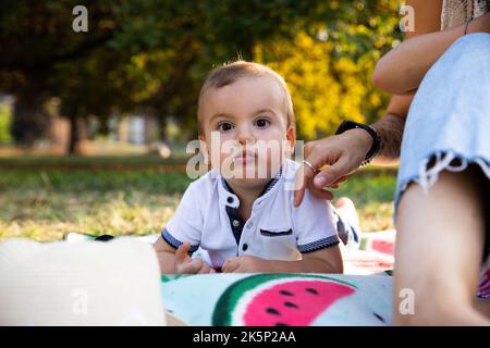 Das Kleinkind, das mit vollem Mund auf einer Picknickdecke liegt, wird von der neben ihm sitzenden Mutter überwacht. Stockfoto