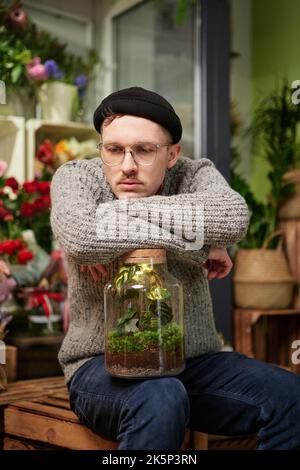 Schöner Gewächshausbesitzer oder Gärtner mit Pflanzen in einem Glas. Attraktive männlich müde Hipster Unternehmer Floristen trägt Pullover, Beanie und Brille im Blumenladen. Hochwertiges vertikales Bild Stockfoto