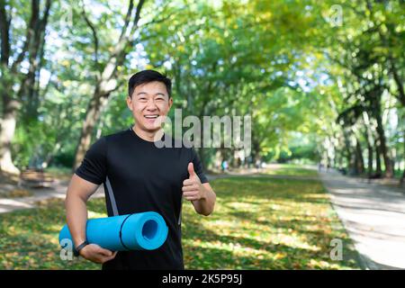 Portrait von Yoga-Trainer, asiatischem Mann, der die Kamera anschaut und lächelt und die Daumen hoch hält, Mann im Herbstpark, der Sportmatte hält, Outdoor-Meditation. Stockfoto