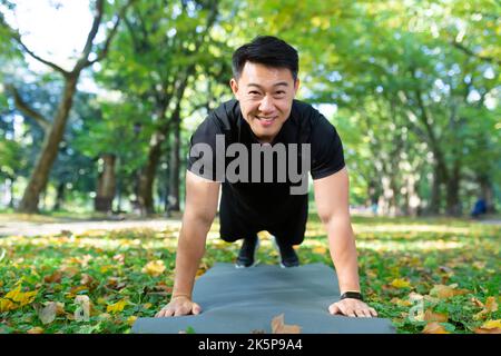 Porträt eines glücklichen männlichen Athleten, eines Asiaten, der die Kamera anschaut und lächelt, indem er an sonnigen Tagen im Herbstpark Liegestütze und Planken macht. Stockfoto