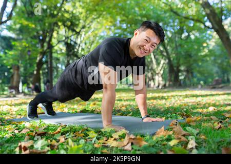 Porträt eines glücklichen männlichen Athleten, eines Asiaten, der die Kamera anschaut und lächelt, indem er an sonnigen Tagen im Herbstpark Liegestütze und Planken macht. Stockfoto