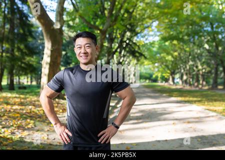 Portrait eines asiatischen Athleten im Herbstpark an sonnigen Tagen, Mann in Sportkleidung lächelnd und schauend auf die Kamera, Fitnesstrainer vor dem Training. Stockfoto