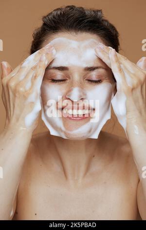 Glückliche Frau mittleren Alters mit schäumenden Gesichtsreiniger Waschgesicht isoliert auf beigem Hintergrund. Stockfoto