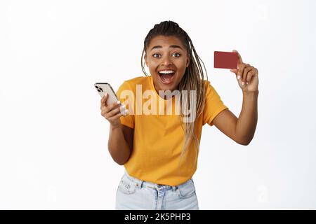 Glückliche junge schwarze Frau zeigt ihre Kreditkarte nach der Verwendung der Handy-App, lacht mit fröhlichem Gesichtsausdruck, weißem Hintergrund Stockfoto