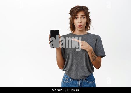 Überrascht und erstaunt junge Frau, zeigte mit dem Finger auf den Smartphone-Bildschirm, stellte Fragen, zeigte smeth auf dem Handy, stand gegen Weiß Stockfoto