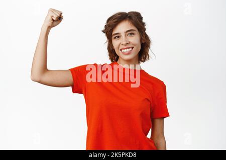 Workout und starke Frau Konzept. Lächelnde junge Frau mit Bizeps, beugenden Muskeln, prahlend mit ihren starken Armen, stehend über weißem Hintergrund Stockfoto