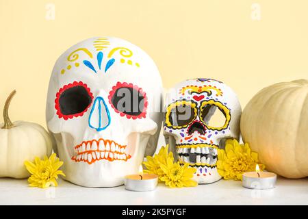 Bemalte menschliche Schädel mit Blumen, Kürbissen und brennenden Kerzen auf dem Tisch in der Nähe der beigen Wand. El Dia de Muertos Stockfoto