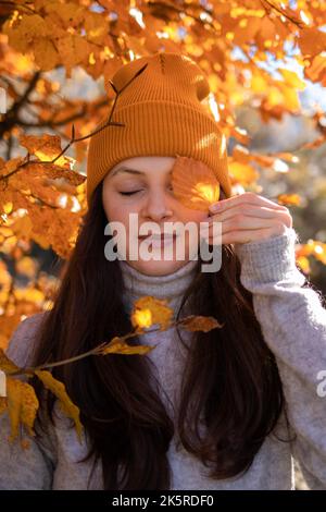 Ein Porträt einer Brünette in einer orangefarbenen Beanie im orangefarbenen Laubbaum. Stockfoto