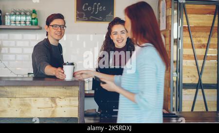 Eine fröhliche Kassiererin verkauft Kaffee zum Mitnehmen an eine junge Frau und akzeptiert Zahlungen, während ein gut aussehender Barista gerade Getränke zubereitet und mitbringt. Stockfoto