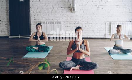 Junge attraktive Yogaschülerinnen meditieren am Ende der Übung in Lotusstellung auf Matten im schönen Lichtstudio. Entspannung Ruhe Balance Konzept. Stockfoto