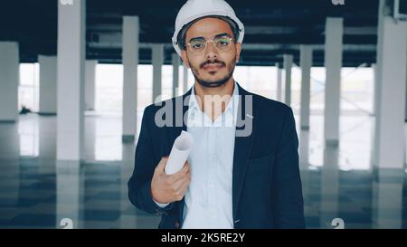 Porträt eines Bauinvestors aus dem Nahen Osten, der Blaupause hält und Helm trägt, der im neuen Werksgebäude steht und die Kamera anschaut Stockfoto