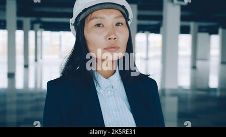 Porträt einer eleganten asiatischen Geschäftsfrau, die Schutzhelm trägt und im leeren Pflanzengebäude steht, das tagsüber zur Miete oder zum Verkauf bereit ist Stockfoto