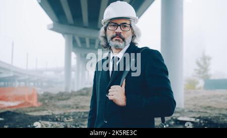 Porträt des Bauprojektleiters, der in Helm und formellem Anzug auf der Baustelle steht und die Kamera anschaut Stockfoto