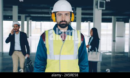 Porträt eines ernsthaften Bauspezialisten mit Sicherheitsuniform, der in einem leeren Gebäude mit Geschäftsleuten im Hintergrund steht. Immobilien- und Reparaturkonzept. Stockfoto